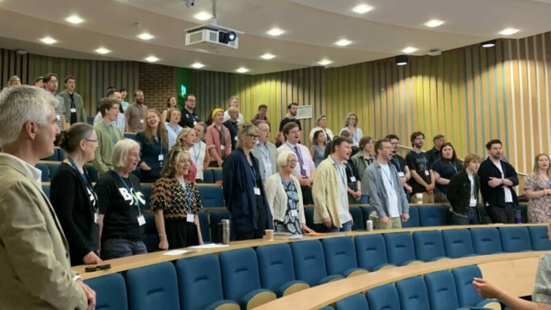 A large group of people stand in a light conference room singing